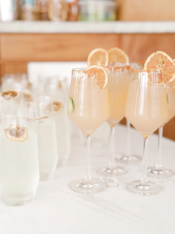 Wedding signature drinks at a wedding cocktail station with stemmed and highball glasses, citrus slices, ice, and salt rims on a bar counter