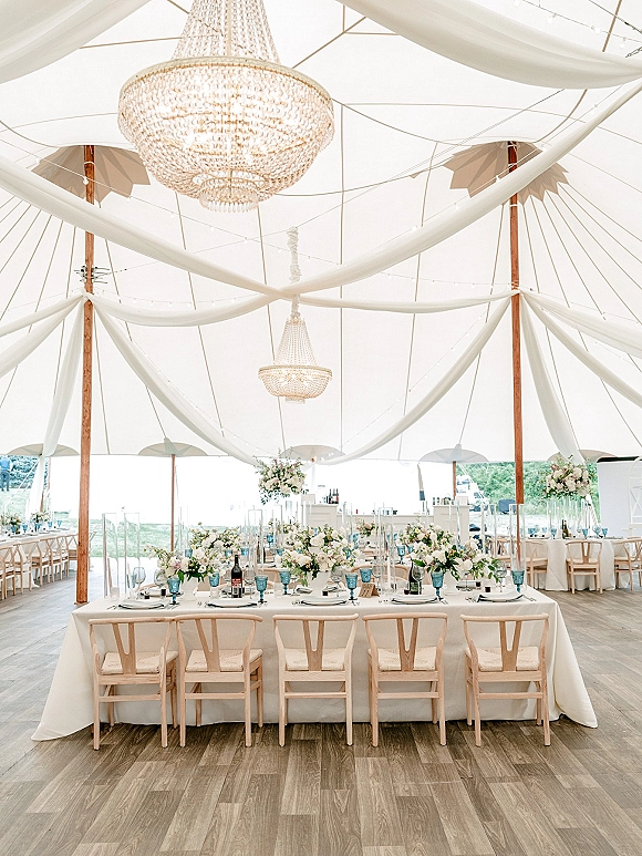 Reception tablescape with sailcloth tent reception draping, blue goblets and taper candles under crystal chandeliers and string lights in a bright tent