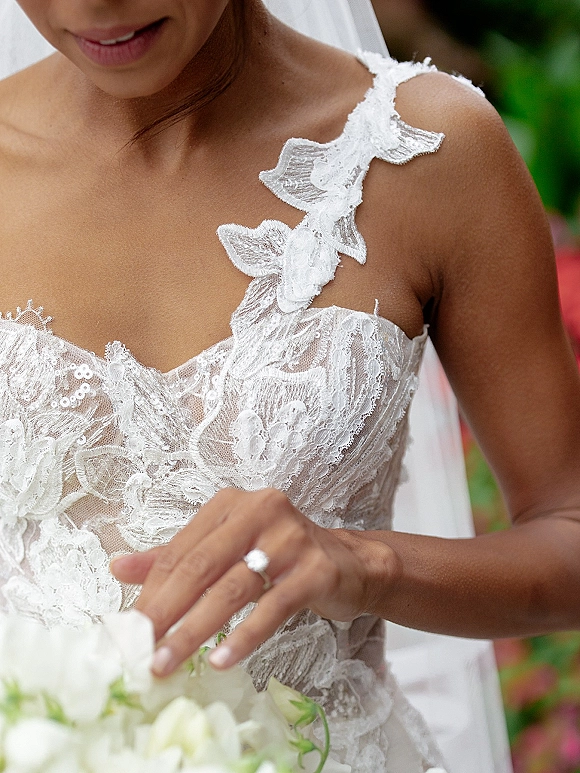 Wedding dress close-up showing a one shoulder wedding dress with lace appliqué, veil and engagement ring while holding a white rose bouquet in a blurred garden
