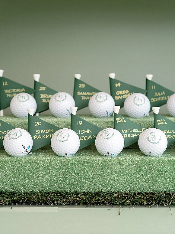 Golf escort cards with golf ball place cards and mini flags, calligraphy names on green felt faux grass atop a tabletop and green wall backdrop