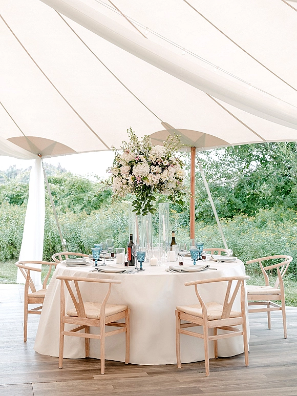 Reception tablescape with round wedding table on white tablecloth, tall floral centerpiece, blue goblets and candles under a white tent canopy