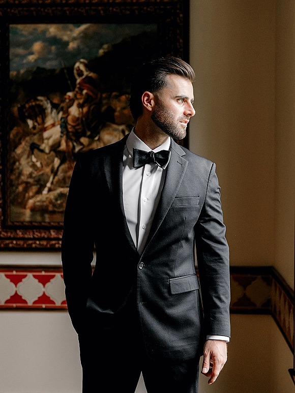 Groom portrait in a black tie tuxedo and bow tie, standing indoors by a stair railing with a framed painting behind him