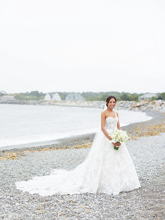 Bridal portrait of a bride holding a white flower bouquet, wearing a lace wedding dress and veil on a pebble beach by the ocean