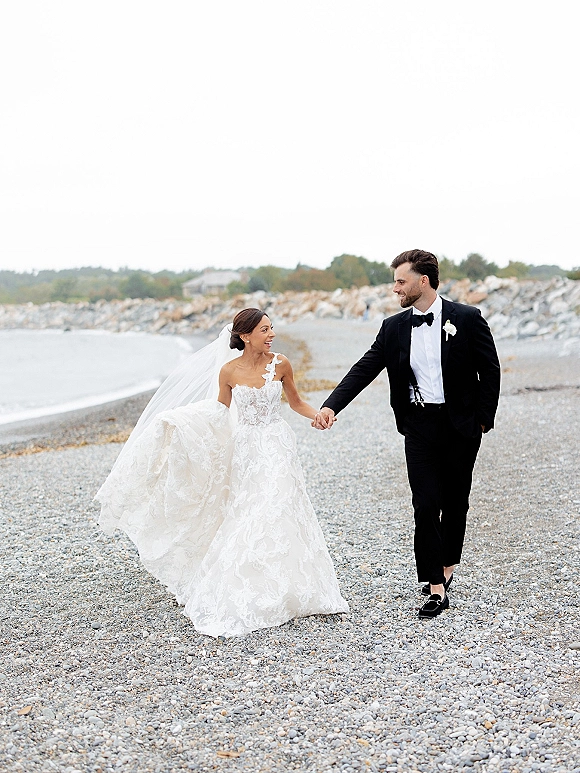 Couple portrait of bride and groom walking hand in hand on a pebble beach, veil in wind, tuxedo and bow tie by ocean rocks under overcast sky