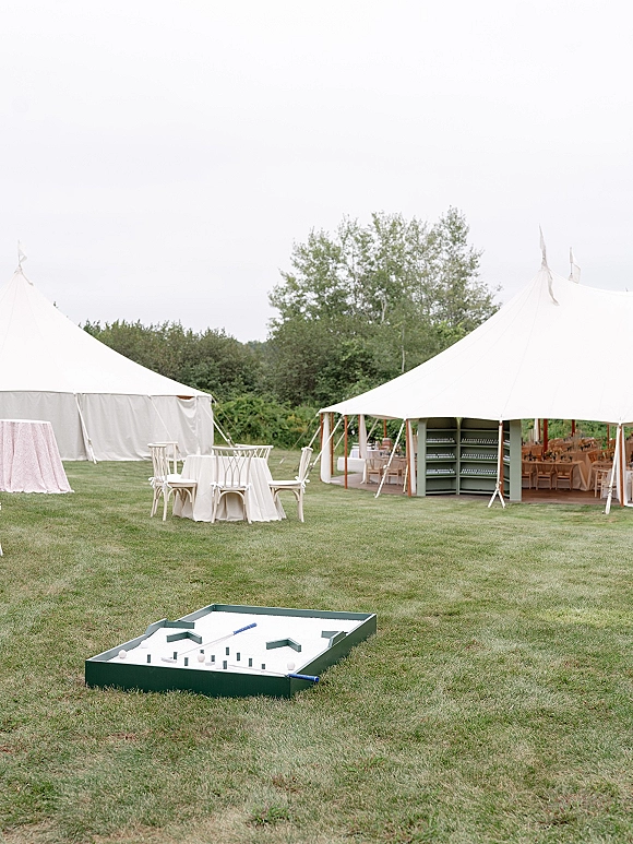 Outdoor wedding lounge with wedding cocktail hour seating under white event tents, round linen tables, wooden chairs, and lawn games on grass