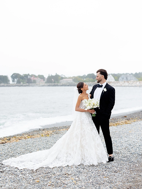 Couple portrait of bride and groom portrait looking at each other on a rocky beach, her veil and long train flowing with bouquet under overcast sky