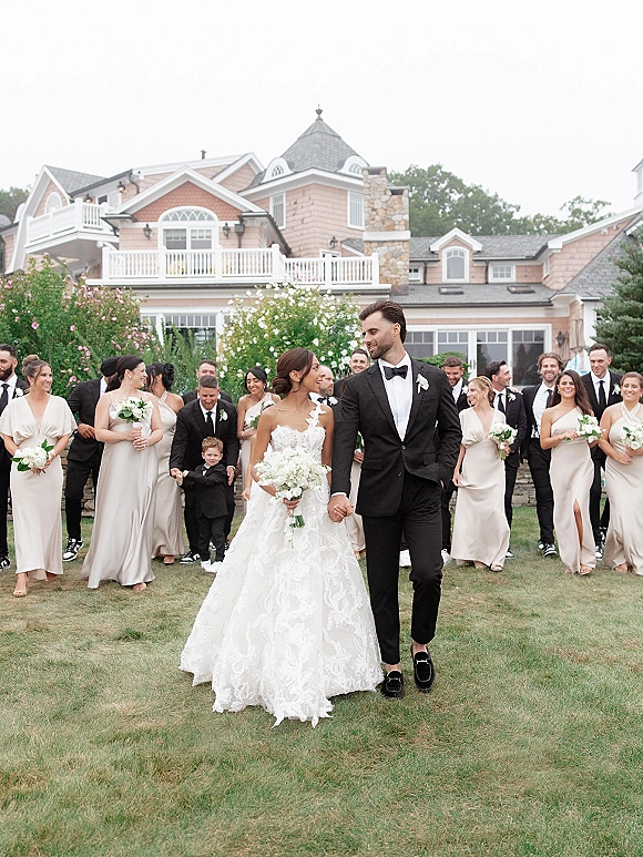 Wedding party portrait of bride and groom holding hands as they walk with bridesmaids and groomsmen on a garden lawn by an estate house