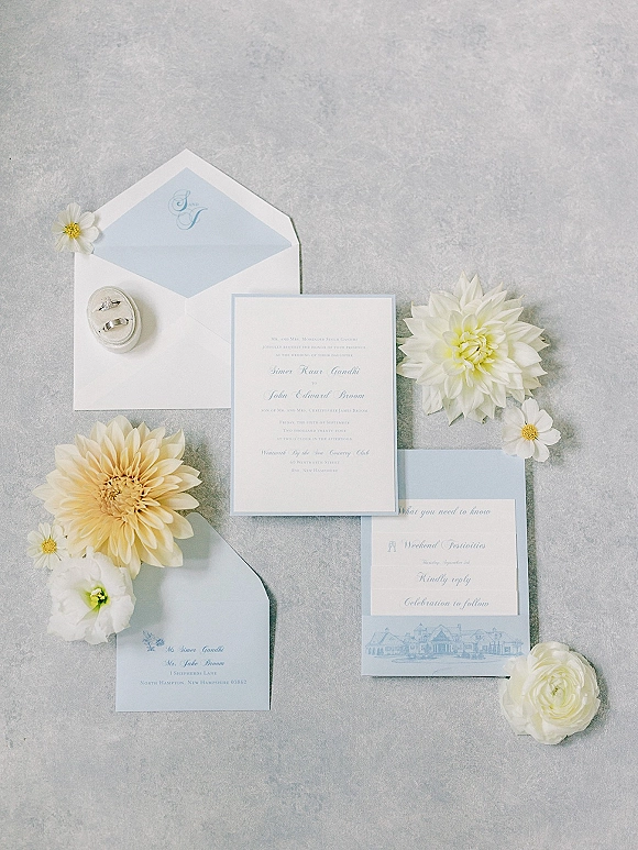 Invitation suite of blue and white wedding invitations with monogram-lined envelopes, wax seal, rings, and dahlias on gray texture