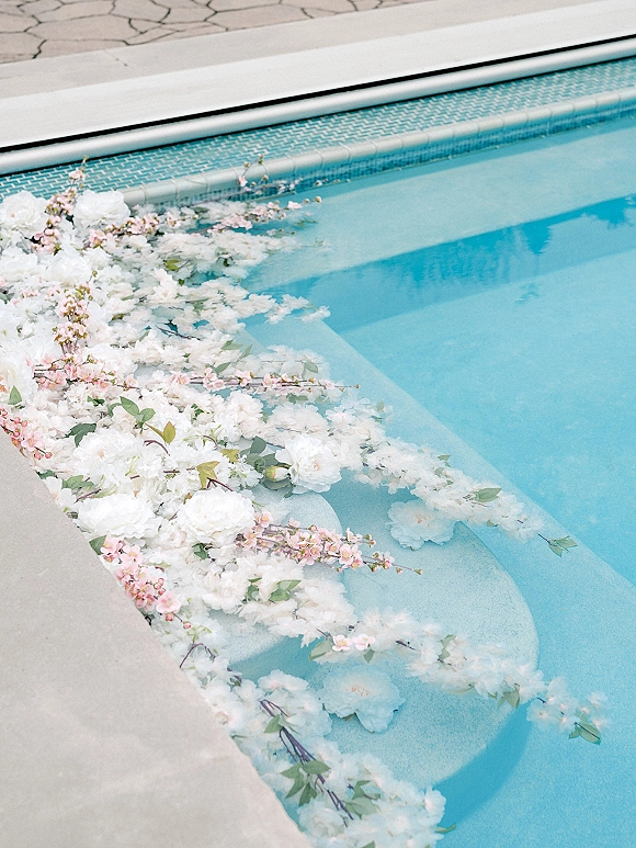 Pool wedding decor with a floating floral arrangement of white blooms, blush blossoms, and greenery garland drifting by the pool steps
