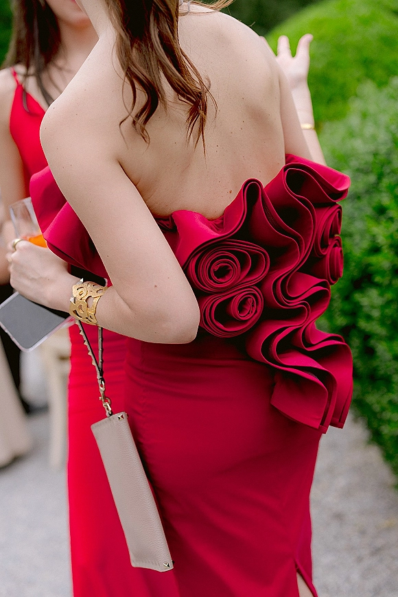 Bridesmaid dress in vibrant red with strapless ruffled detail, styled with a gold cuff bracelet and clutch on an outdoor walkway by a green hedge