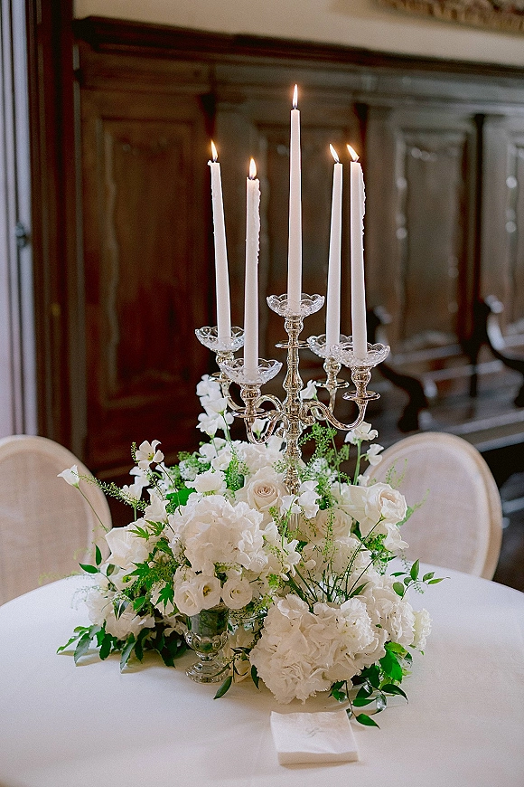 Wedding centerpiece with a silver candelabra, taper candles, and white roses and hydrangeas on a round linen table against wood paneling