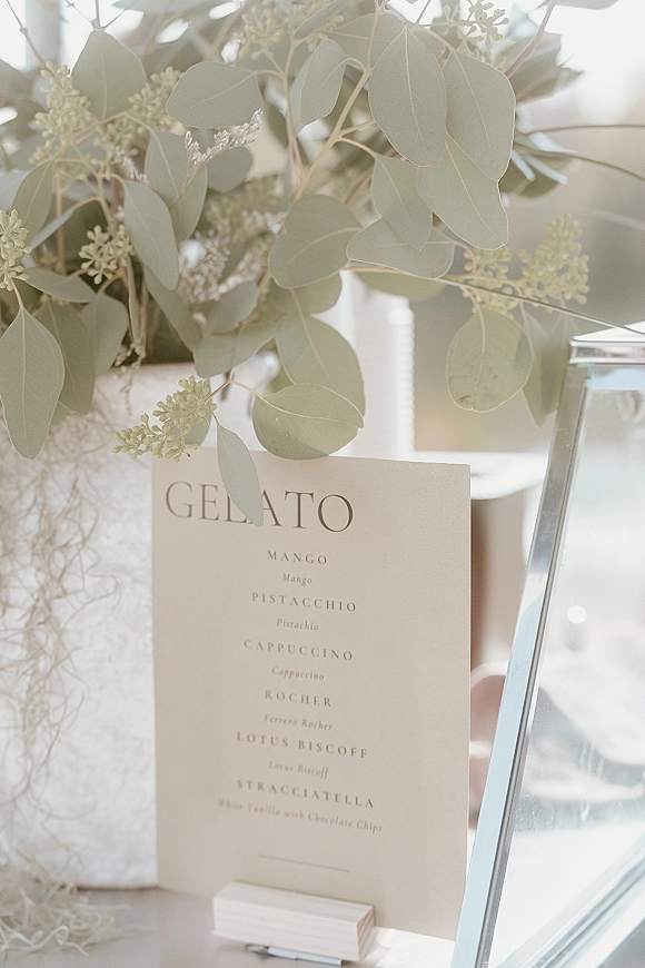 Gelato menu sign for a wedding gelato bar in a wooden holder, printed card beside a white vase with eucalyptus on a sunlit tabletop