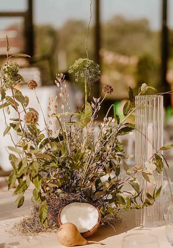 Wedding centerpiece with greenery arrangement in a clear ribbed glass vase, featuring wildflowers, dried florals, pear, coconut, and moss on linen tablecloth