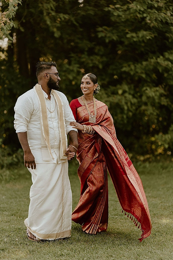 Couple portrait of a South Indian wedding couple holding hands on a garden lawn, bride in red silk saree with gold jewelry and bindi