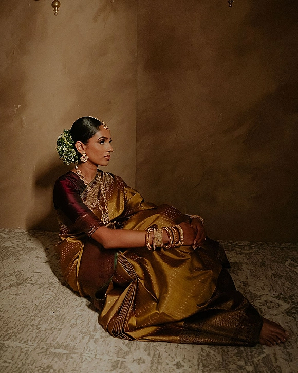 Bridal portrait of a South Indian bride in a silk saree with gold jewelry and jasmine gajra, seated by a textured plaster wall