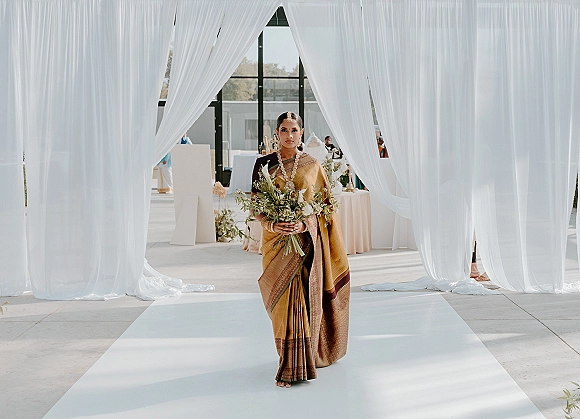 Bridal portrait of a South Asian bride holding a white and green bouquet, wearing a gold silk sari and jewelry in a draped indoor aisle setting