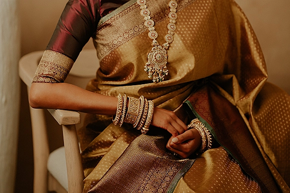 Bridal jewelry on a gold silk saree, featuring a statement necklace, pendant, and stacked bangles beside an indoor chair and wall