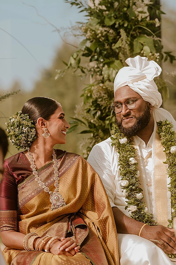 Couple portrait of a South Indian wedding couple seated, gazing at each other in garlands beneath a floral arch with greenery behind