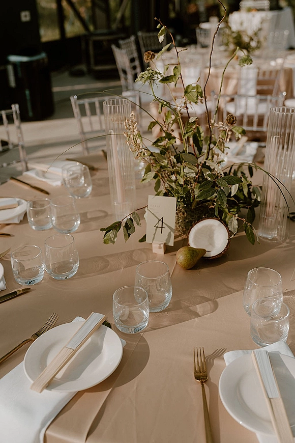 Reception tablescape with a wedding table centerpiece of greenery, white taper candles in ribbed holders, gold flatware, and fruit in window light