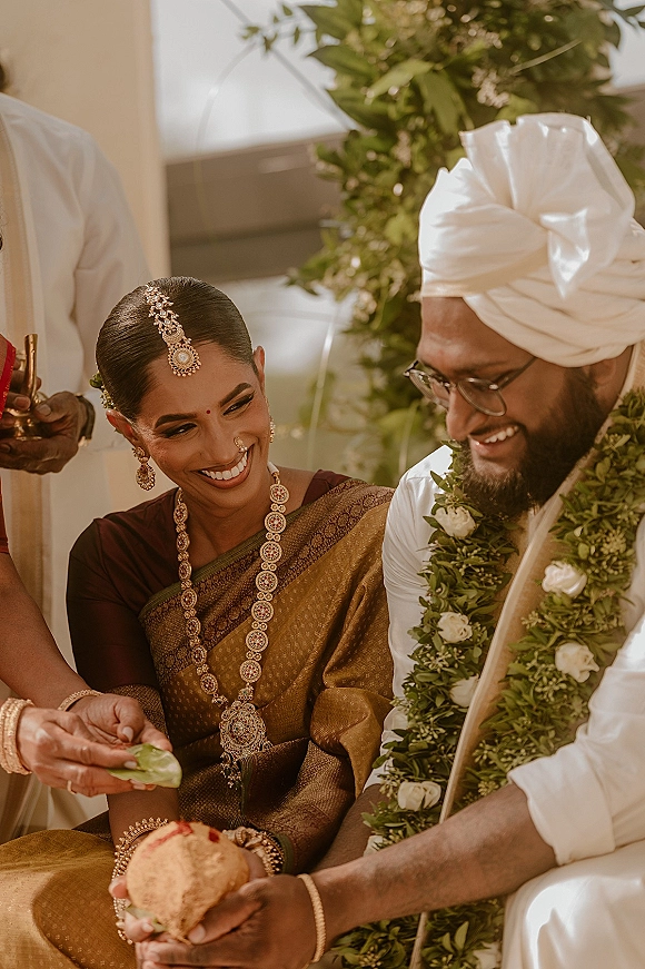 Hindu wedding ceremony with bride in silk saree and groom in white turban exchanging garlands, coconut ritual before greenery backdrop indoors