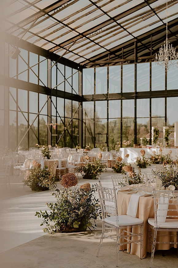 Wedding reception decor with round tables, beige linens, clear acrylic chairs, and floral greenery centerpieces beneath chandeliers in a glass greenhouse