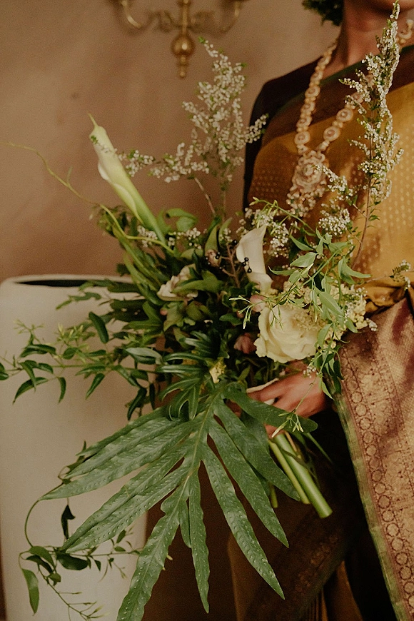Bridal bouquet of calla lilies and white roses with lush greenery and a monstera leaf, held by a bride in a sari indoors under a chandelier