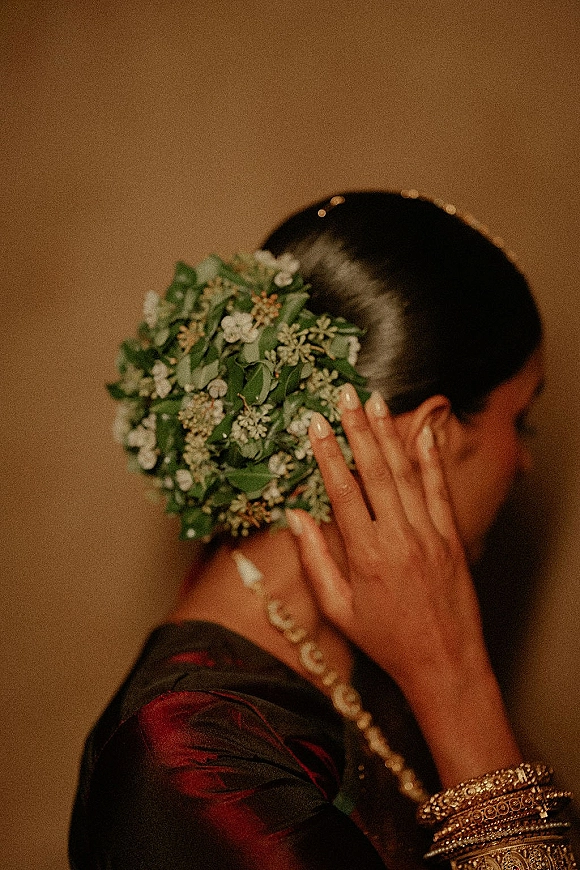 Bridal hair flowers frame a low bun with white blooms and greenery hairpiece, with mehndi hands and gold bangles by a neutral wall
