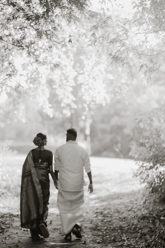 Couple walking away holding hands, bride and groom walking in saree and traditional attire under a sunlit tree canopy path