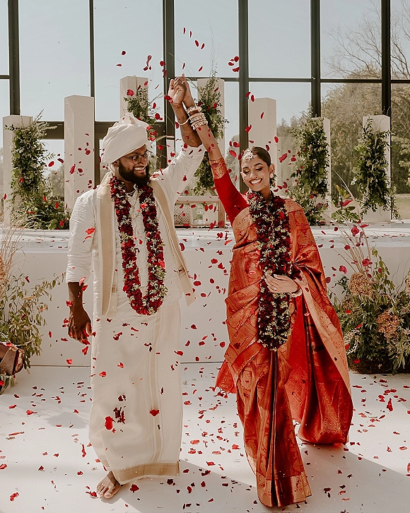 Wedding recessional with a Sikh wedding couple holding hands as red rose petals fall, bride in red sari and groom in turban by glass windows