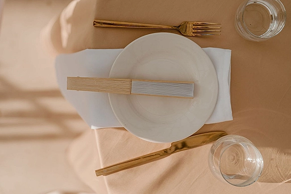 Reception tablescape with wedding place setting featuring a white plate, menu card, gold flatware, glass tumbler on beige tablecloth