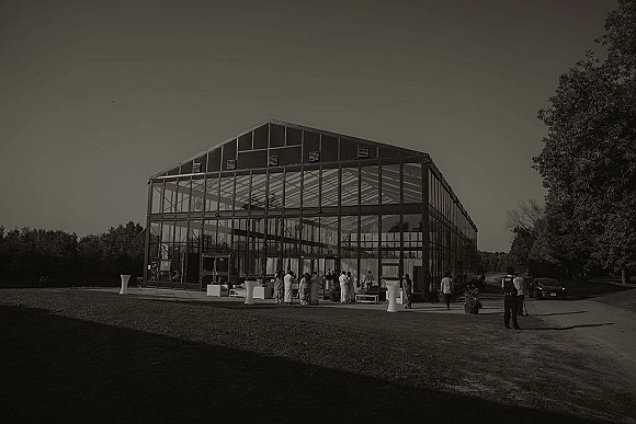 Wedding reception venue with a glass greenhouse structure, cocktail tables and white lounge seating set on a lawn with trees behind