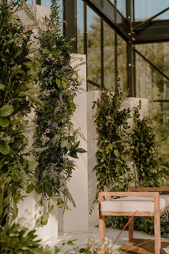Wedding ceremony backdrop with greenery garlands and white floral accents on white plinths, set before a glass wall with trees beyond.
