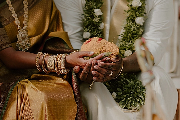 Wedding ritual hands in a hindu wedding ritual holding a coconut with betel leaves and gold bangles, seated couple blurred behind