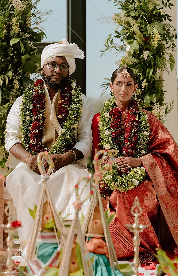 Couple portrait of a south indian wedding couple seated on a stage, wearing floral garlands as the bride holds a bouquet against a greenery wall