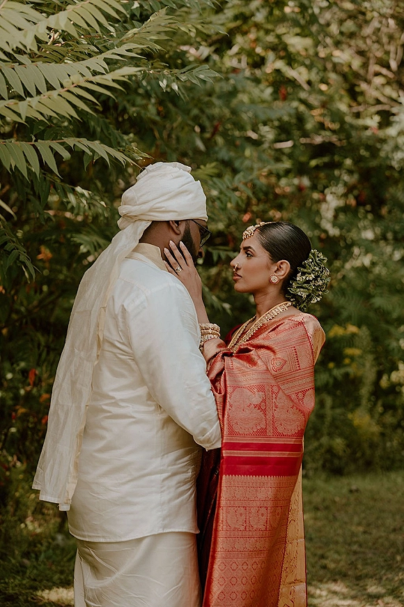 Couple portrait of a South Asian wedding couple as the bride in a red saree touches the groom’s face, garden greenery behind them