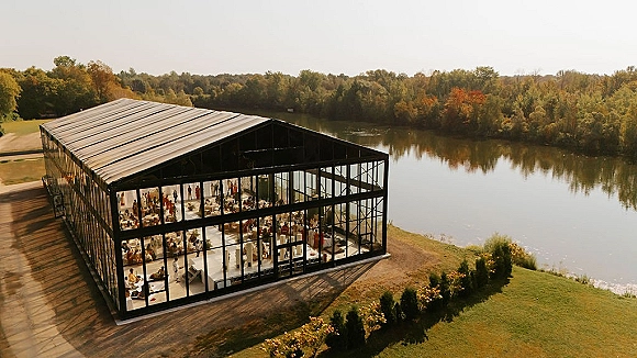 Glass wedding venue with glass walls and black metal frame set for reception, overlooking a lakeside lawn with trees and sky beyond