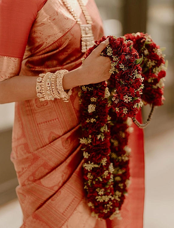 Wedding garland with red wedding garland blooms and jasmine flowers, held by a bride in saree with bangles against a soft indoor blur
