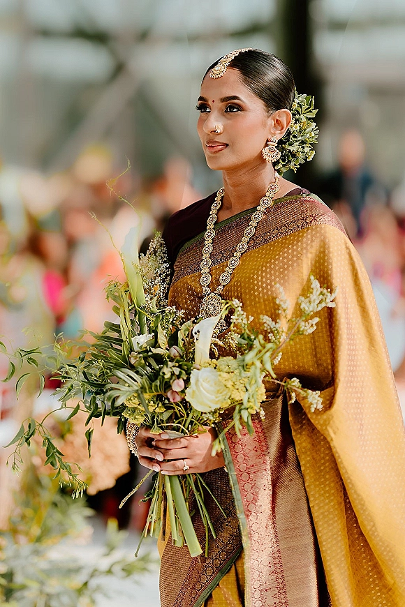 Bridal portrait of an Indian bride portrait in a silk saree with gold jewelry and bouquet, greenery behind with blurred guests