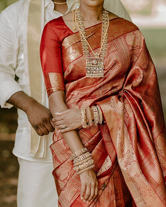Bride portrait in a red silk saree with gold jewelry and bangles, groom behind her, framed by soft natural light and greenery outdoors
