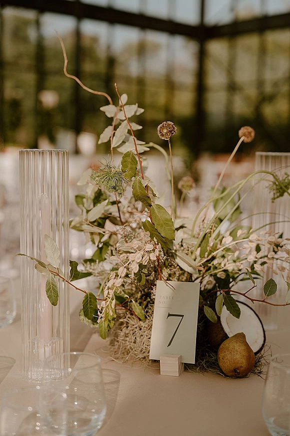 Reception tablescape with wedding table centerpiece in ribbed glass vases, greenery, fruit, and table number card in a glass greenhouse setting