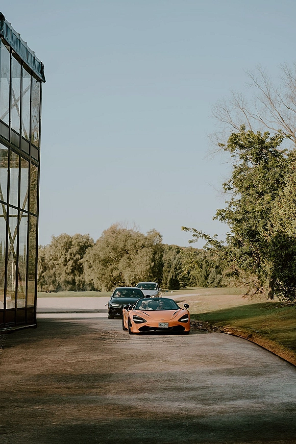 Wedding car exit with a convertible sports car as the wedding getaway car pulls down a venue driveway beside a glass building and trees