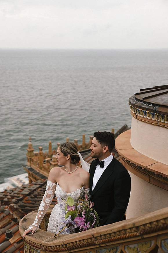 Couple portrait of bride and groom leaning on a stone rooftop railing, bride with purple bouquet and veil, ocean behind under overcast sky