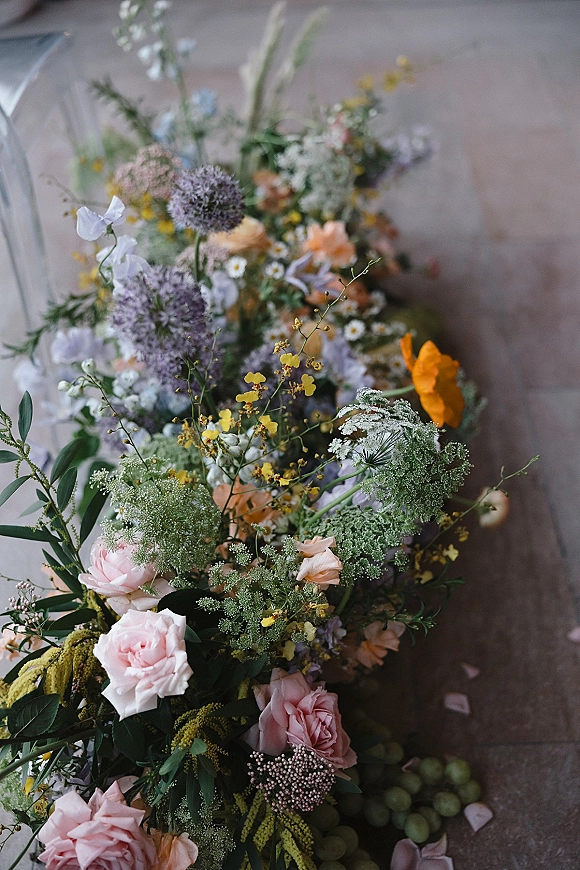 Wedding floral arrangement with ceremony aisle flowers in pastel roses, wildflowers, and greenery on a wood floor beside a clear chair