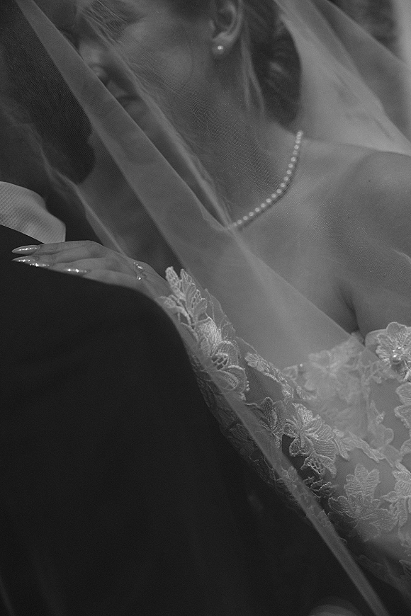 Wedding kiss portrait of bride and groom under a veil, her lace dress and pearl necklace visible against a dark backdrop