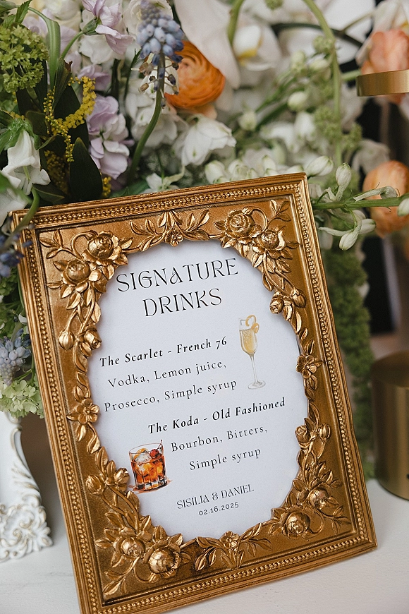 Signature drinks sign in an ornate gold frame on a white tablecloth with floral accents and greenery beside a blurred bar area