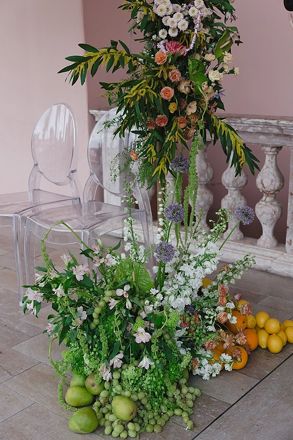 Wedding floral installation with ceremony floral pillar, hanging blooms and greenery, plus fruit accents by a stone balcony and pink wall