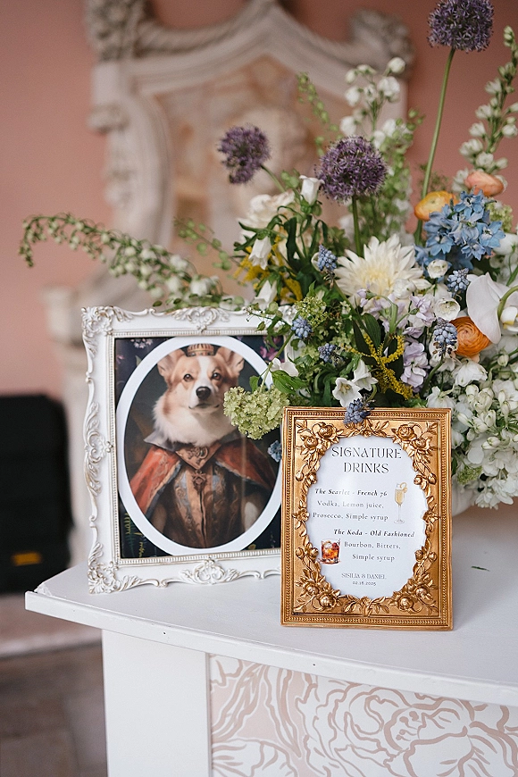 Signature drinks sign in an ornate gold frame with signature cocktail menu, paired with blue and yellow florals on a pedestal table by a pink wall