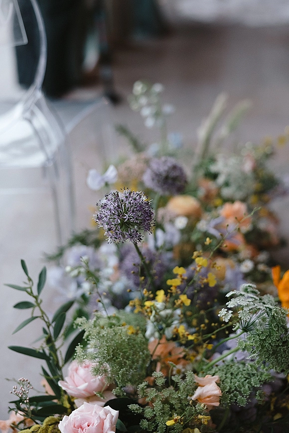 Wedding florals wildflower wedding centerpiece with purple allium, pastel roses and greenery beside an acrylic chair in a blurred indoor reception space