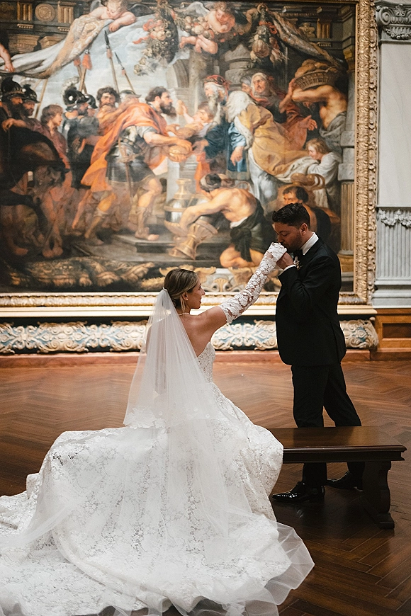 Couple portrait of groom kissing bride’s gloved hand as she sits in a lace gown and veil on a bench before a grand painting