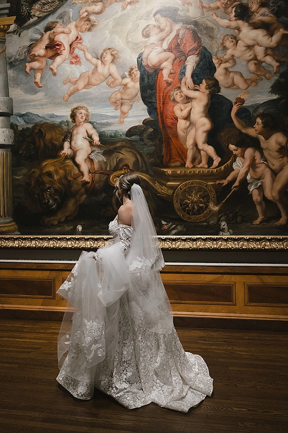 Bridal portrait of a bride with cathedral veil, showing her lace gown long train as she faces a gilded painting in a museum gallery